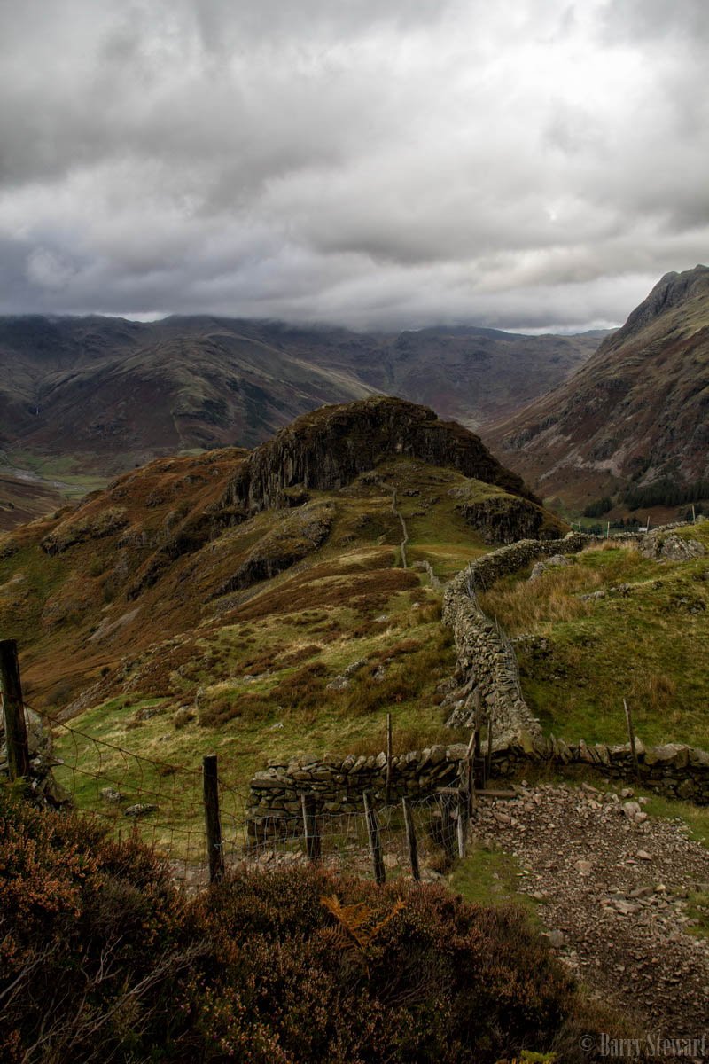 A winding stone wall follows a rugged path through hilly terrain under a cloudy sky, with mountains in the distance.