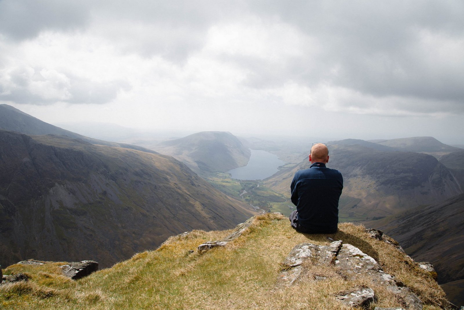 Lake District A person sits on a grassy cliff edge overlooking a valley with mountains and a lake under a cloudy sky.
