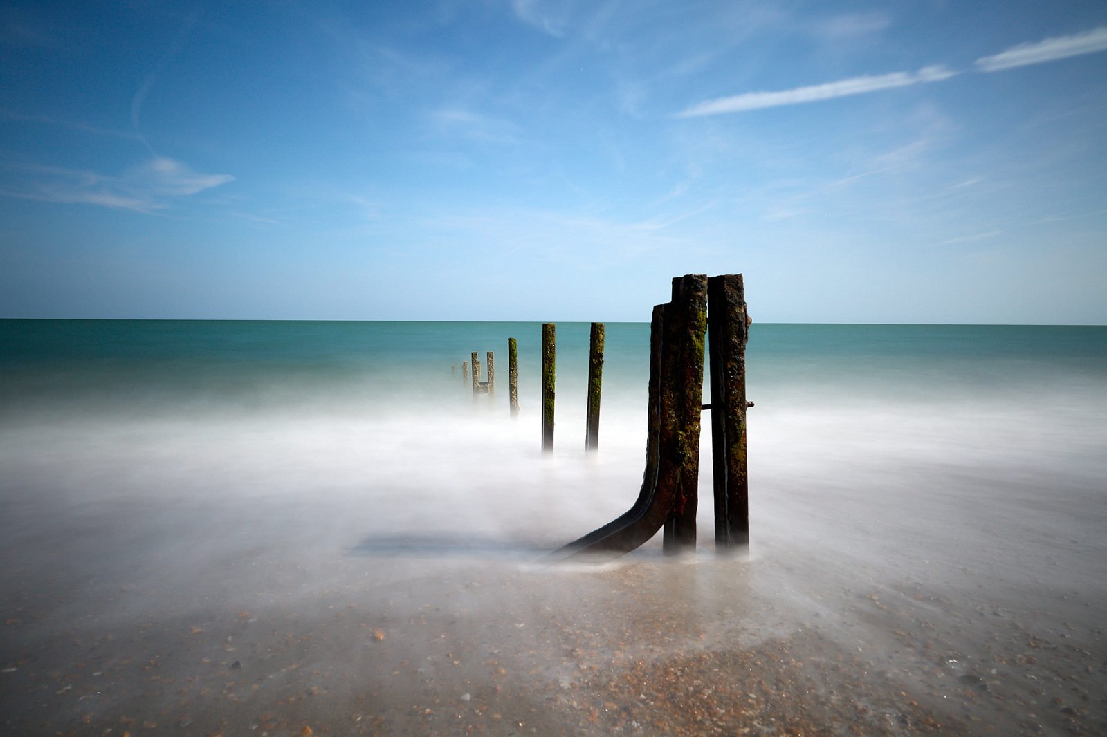 959A4685 Wooden posts extend from the sandy shore into the sea under a blue sky, with waves creating a misty effect around the base of the posts.
