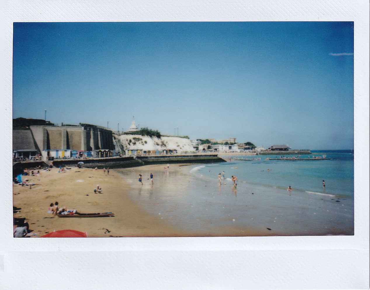 IMG_20190802_0007 Polaroid photo of a sandy beach with people sunbathing and swimming; coastal cliffs, buildings, and a clear blue sky in the background.