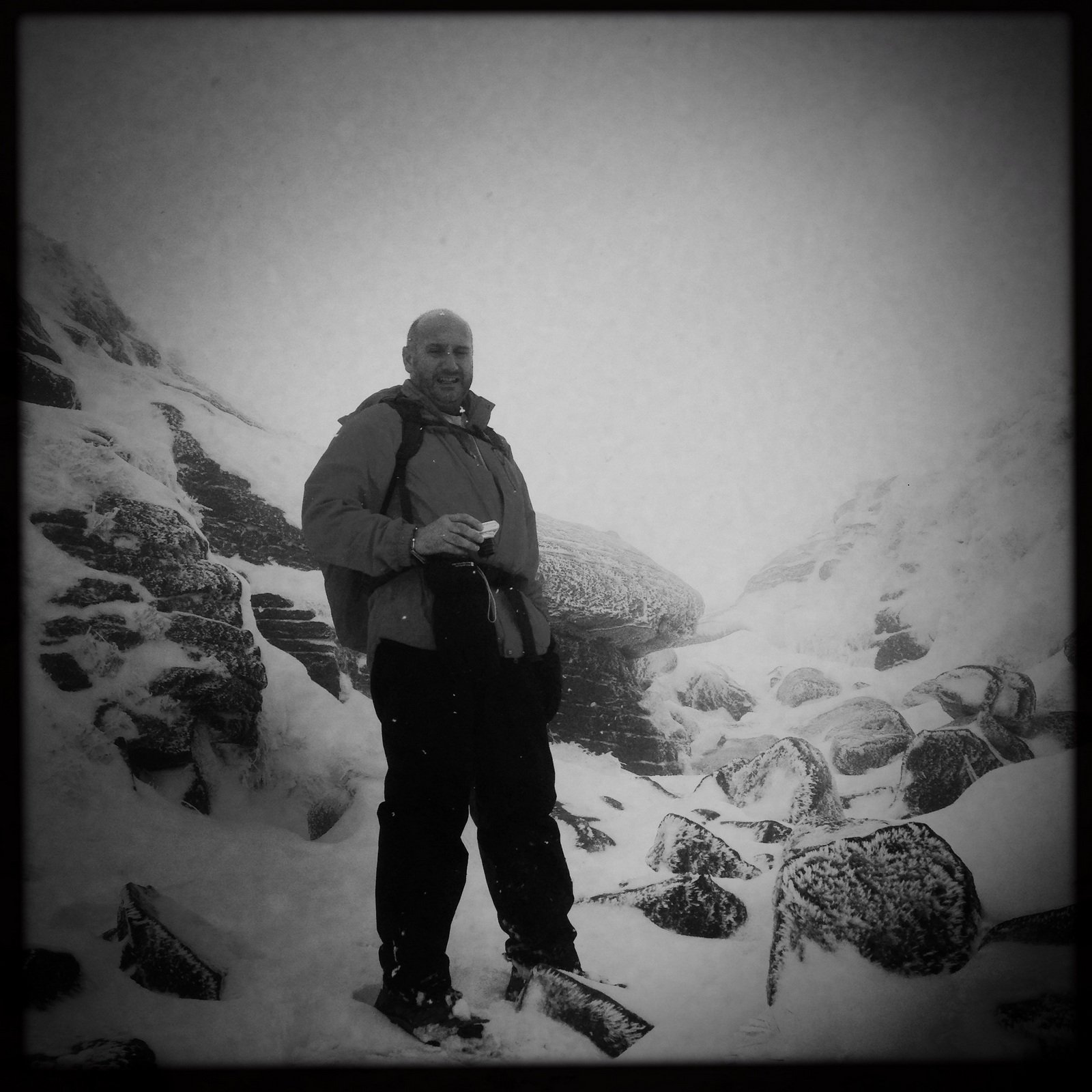 A person stands on a snowy, rocky mountain slope, wearing a jacket and holding an object in one hand. The scene is foggy and wintry.
