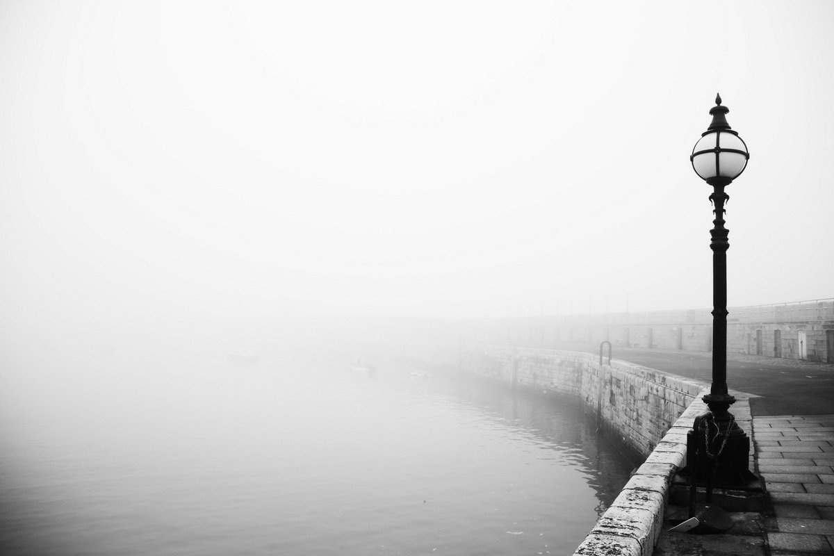 A streetlamp stands on a stone pier beside calm water, with thick fog obscuring most of the background and distant objects.